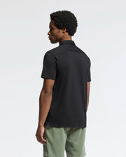 A person with short curly hair, slightly turned away, wears the Psycho Bunny MENS BRYAN CAMP COLLAR SHORT SLEEVE SHIRT in black and olive green shorts. The light background emphasizes their outfit's moisture-wicking fabric.