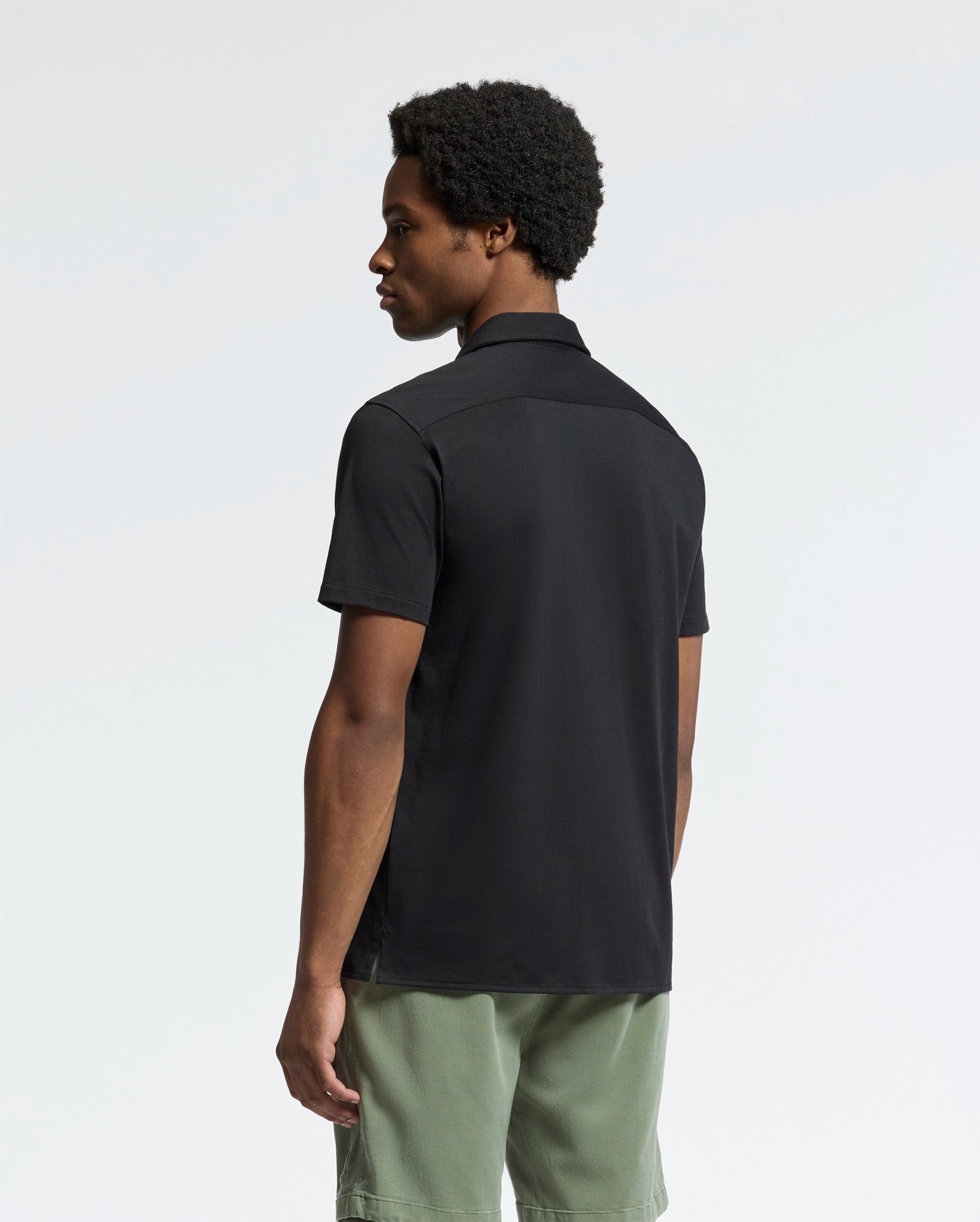 A person with short curly hair, slightly turned away, wears the Psycho Bunny MENS BRYAN CAMP COLLAR SHORT SLEEVE SHIRT in black and olive green shorts. The light background emphasizes their outfit's moisture-wicking fabric.