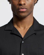 A person is wearing the Psycho Bunny MENS BRYAN CAMP COLLAR SHORT SLEEVE SHIRT (B6Q581F200), which is black and features a small embroidered logo on the chest. The image highlights their upper body against a plain, light-colored background, with their face partially cropped.