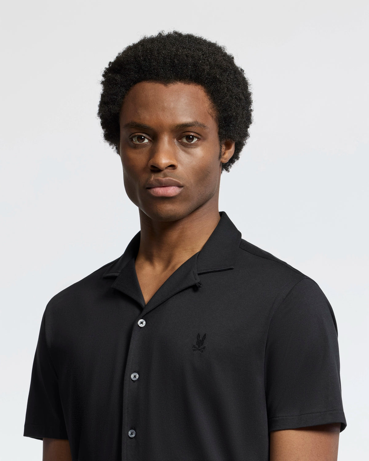 A person with short, curly hair wearing a black Psycho Bunny MENS BRYAN CAMP COLLAR SHORT SLEEVE SHIRT, made from moisture-wicking Pima cotton, stands against a plain background with a neutral expression.