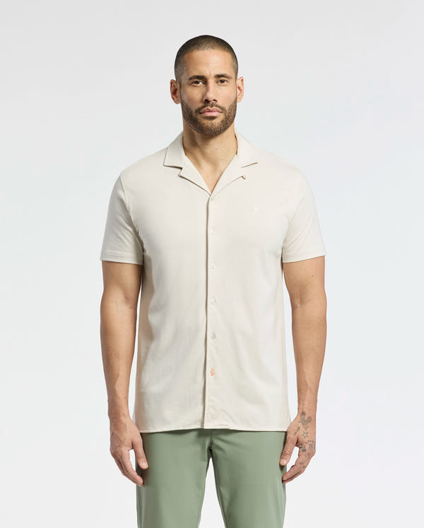 A man with short hair and a beard is wearing a Psycho Bunny MENS BRYAN CAMP COLLAR SHORT SLEEVE SHIRT in light beige and green pants. He stands against a white background, looking directly at the camera with his arms by his sides.