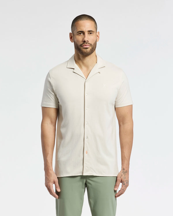 A man with short hair and a beard is wearing a Psycho Bunny MENS BRYAN CAMP COLLAR SHORT SLEEVE SHIRT in light beige and green pants. He stands against a white background, looking directly at the camera with his arms by his sides.