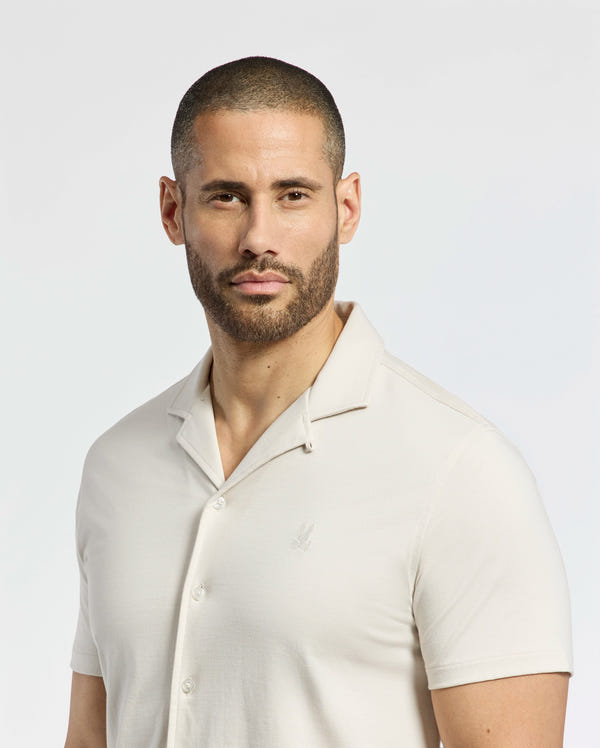 A man with a neat beard and short hair wears a light-colored Psycho Bunny MENS BRYAN CAMP COLLAR SHORT SLEEVE SHIRT - B6Q581F200, standing against a plain background.