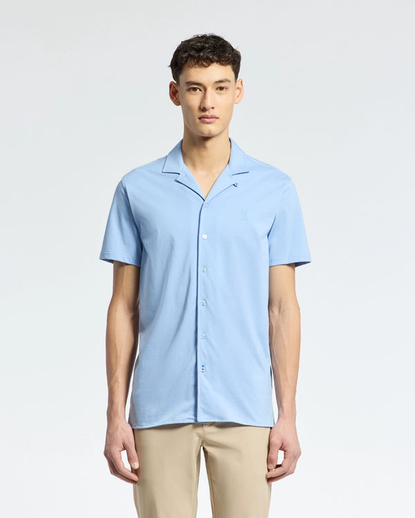 A man wearing Psycho Bunny's light blue MENS BRYAN CAMP COLLAR SHORT SLEEVE SHIRT, made of moisture-wicking performance fabric, and beige pants stands against a white background. Facing the camera with a neutral expression, he embodies effortless style and comfort.