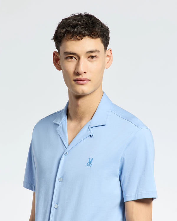 A person with short, dark hair is wearing a Psycho Bunny MENS BRYAN CAMP COLLAR SHORT SLEEVE SHIRT (B6Q581F200) in light blue. The moisture-wicking shirt has an embroidered logo on the chest. They stand against a plain, light background, looking directly at the camera.