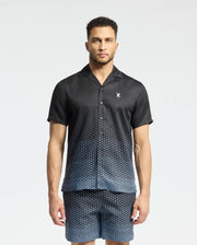 A man wears a Psycho Bunny MENS BENDIGO TENCEL SHIRT - B6Q627F200, featuring short sleeves, a geometric pattern, camp collar, and a white chest logo. He's paired it with matching shorts against a plain background.