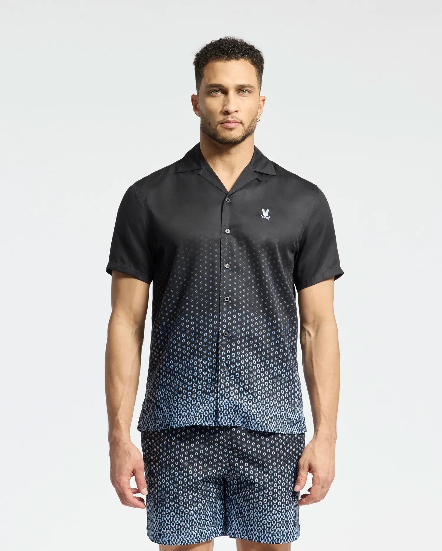 A man wears a Psycho Bunny MENS BENDIGO TENCEL SHIRT - B6Q627F200, featuring short sleeves, a geometric pattern, camp collar, and a white chest logo. He's paired it with matching shorts against a plain background.