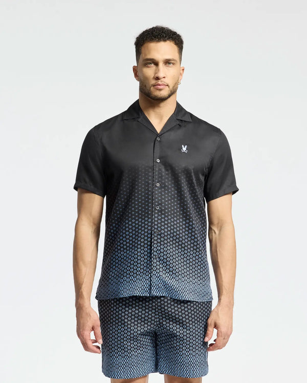 A man wears a Psycho Bunny MENS BENDIGO TENCEL SHIRT - B6Q627F200, featuring short sleeves, a geometric pattern, camp collar, and a white chest logo. He's paired it with matching shorts against a plain background.