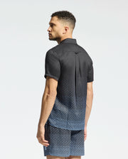 A man stands with his back to the camera, wearing the Psycho Bunny MENS BENDIGO TENCEL SHIRT, a black short-sleeve shirt with blue geometric patterns and a stylish camp collar. It matches his shorts and complements his curly hair and slight beard against a plain white background.