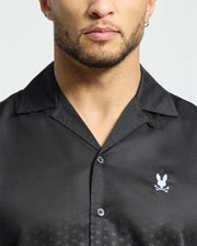 A person with facial hair and an earring is wearing the Psycho Bunny MENS BENDIGO TENCEL SHIRT, featuring an ombre black design with a white rabbit and crossed bones symbol on the left side, plus a subtle dotted pattern near the bottom.