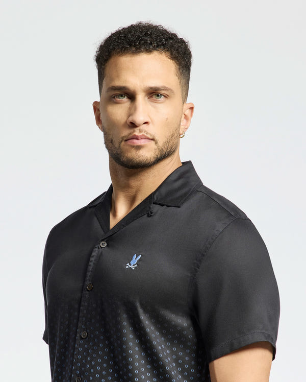 A man with short curly hair and a beard wears the black ombre Psycho Bunny Men's Bendigo Tencel Shirt (B6Q627F200) featuring a camp collar, subtle dotted pattern, and small embroidered emblem. He's turned slightly to his right against a plain background.