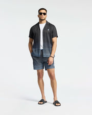 A man wears Psycho Bunny's MENS BENDIGO TENCEL SHIRT, a black and white ombre camp collar shirt over a white T-shirt, paired with matching geometric pattern shorts, black sunglasses, and sandals against a plain white background.