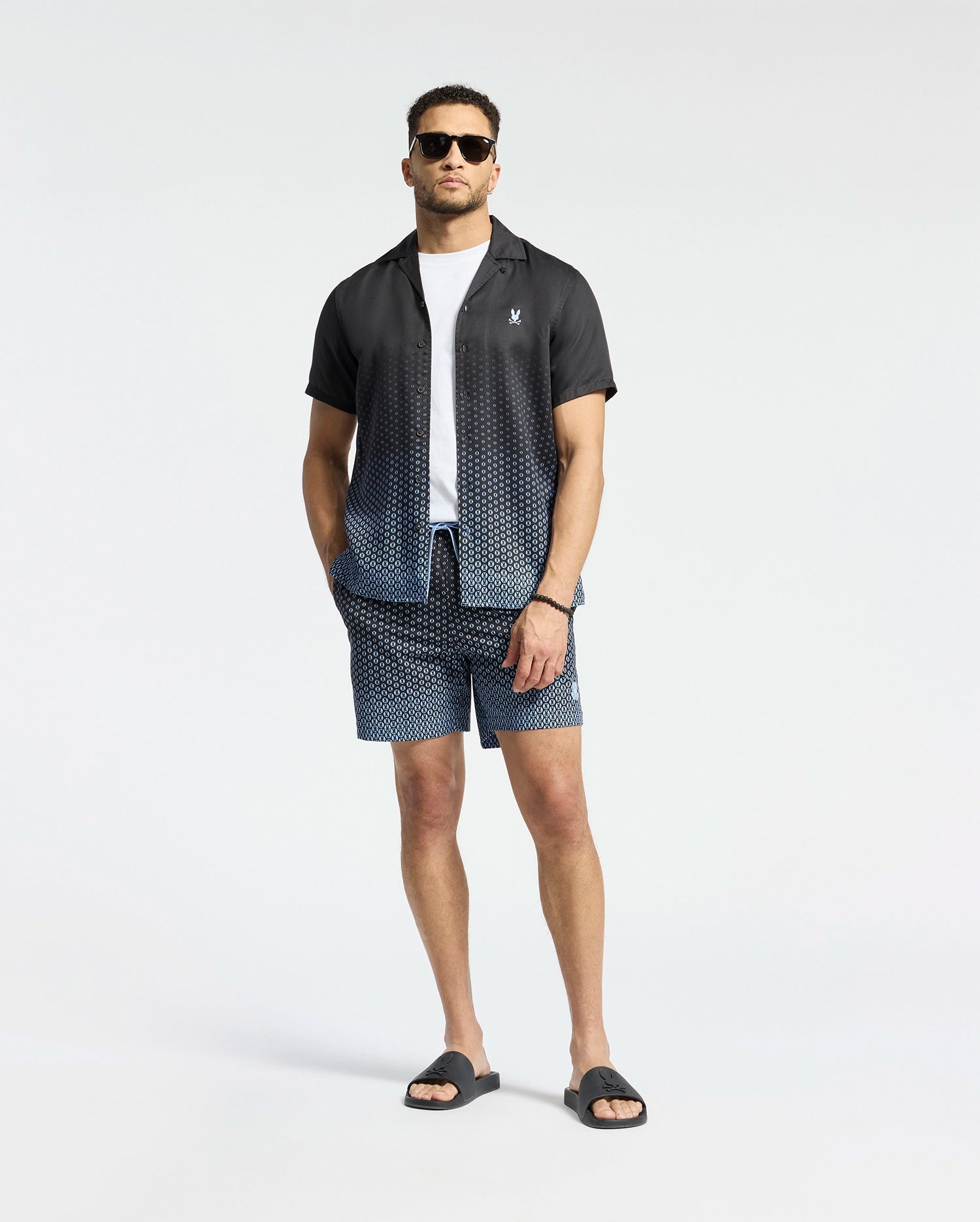 A man wears Psycho Bunny's MENS BENDIGO TENCEL SHIRT, a black and white ombre camp collar shirt over a white T-shirt, paired with matching geometric pattern shorts, black sunglasses, and sandals against a plain white background.