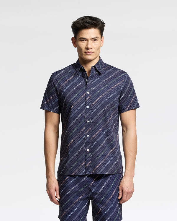 A person stands confidently in a Psycho Bunny MENS RENMARK STRETCH POPLIN WOVEN SHIRT - B6Q629F200, featuring navy-blue stripes on a short-sleeved design, paired with matching pants. The diagonal stripes bring flair against the plain white background.