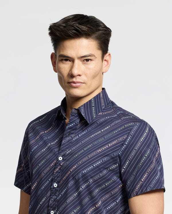 A person with short, dark hair wears a Psycho Bunny MENS RENMARK STRETCH POPLIN WOVEN SHIRT in navy blue. The shirt features multicolored diagonal stripes and text patterns, and has short sleeves against a light background as the person looks toward the camera.