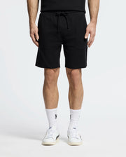 Dressed in Psycho Bunny's MENS TAYLOR LIGHT PIQUE SWEATSHORT - B6R344F200, someone stands against a white backdrop. They pair the black drawstring shorts with logo-emblazoned white socks and white sneakers, showcasing a casual-chic style with only the lower half of their body visible.