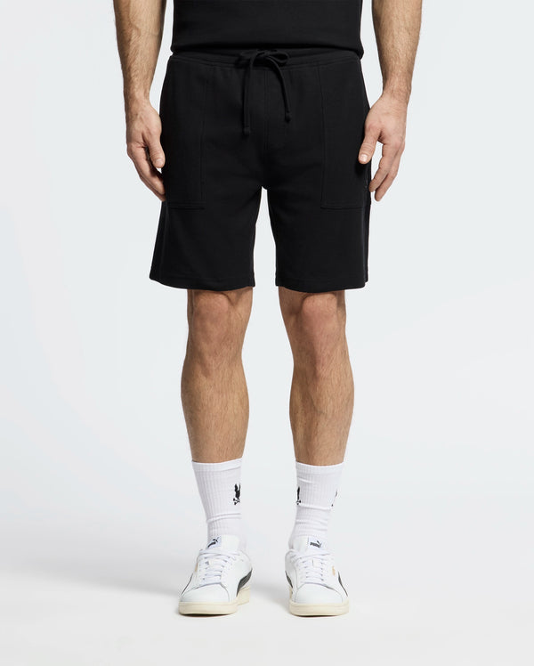Dressed in Psycho Bunny's MENS TAYLOR LIGHT PIQUE SWEATSHORT - B6R344F200, someone stands against a white backdrop. They pair the black drawstring shorts with logo-emblazoned white socks and white sneakers, showcasing a casual-chic style with only the lower half of their body visible.