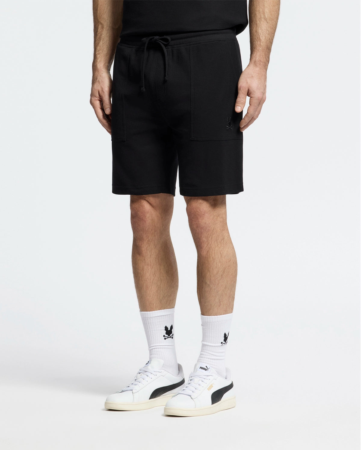 A person strikes a casual-chic pose in Psycho Bunny's MENS TAYLOR LIGHT PIQUE SWEATSHORT - B6R344F200, paired with a black top. White socks featuring black graphics and white sneakers with bold detailing add to their effortless style, all set against a plain white background.