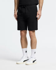 A person strikes a casual-chic pose in Psycho Bunny's MENS TAYLOR LIGHT PIQUE SWEATSHORT - B6R344F200, paired with a black top. White socks featuring black graphics and white sneakers with bold detailing add to their effortless style, all set against a plain white background.