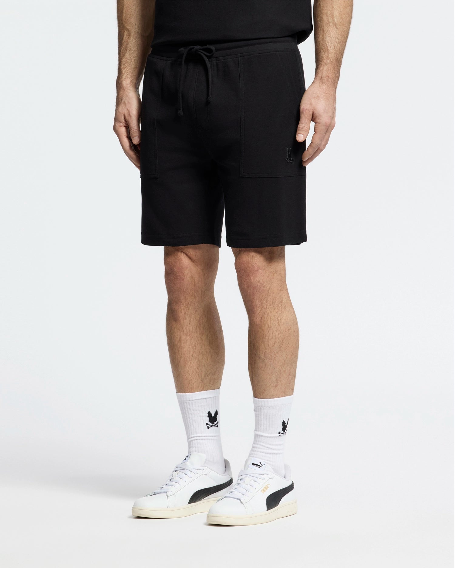 A person strikes a casual-chic pose in Psycho Bunny's MENS TAYLOR LIGHT PIQUE SWEATSHORT - B6R344F200, paired with a black top. White socks featuring black graphics and white sneakers with bold detailing add to their effortless style, all set against a plain white background.