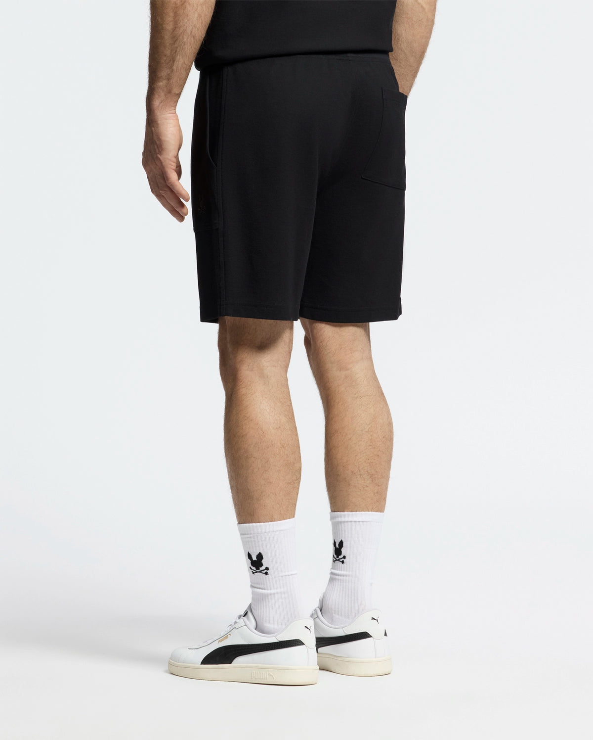 Dressed in a lightweight, black Pima cotton t-shirt and MENS TAYLOR LIGHT PIQUE SWEATSHORT by Psycho Bunny, they pair their outfit with white socks accented by a black logo and chic white sneakers featuring black details against a plain white backdrop, exuding effortless casual-chic style.