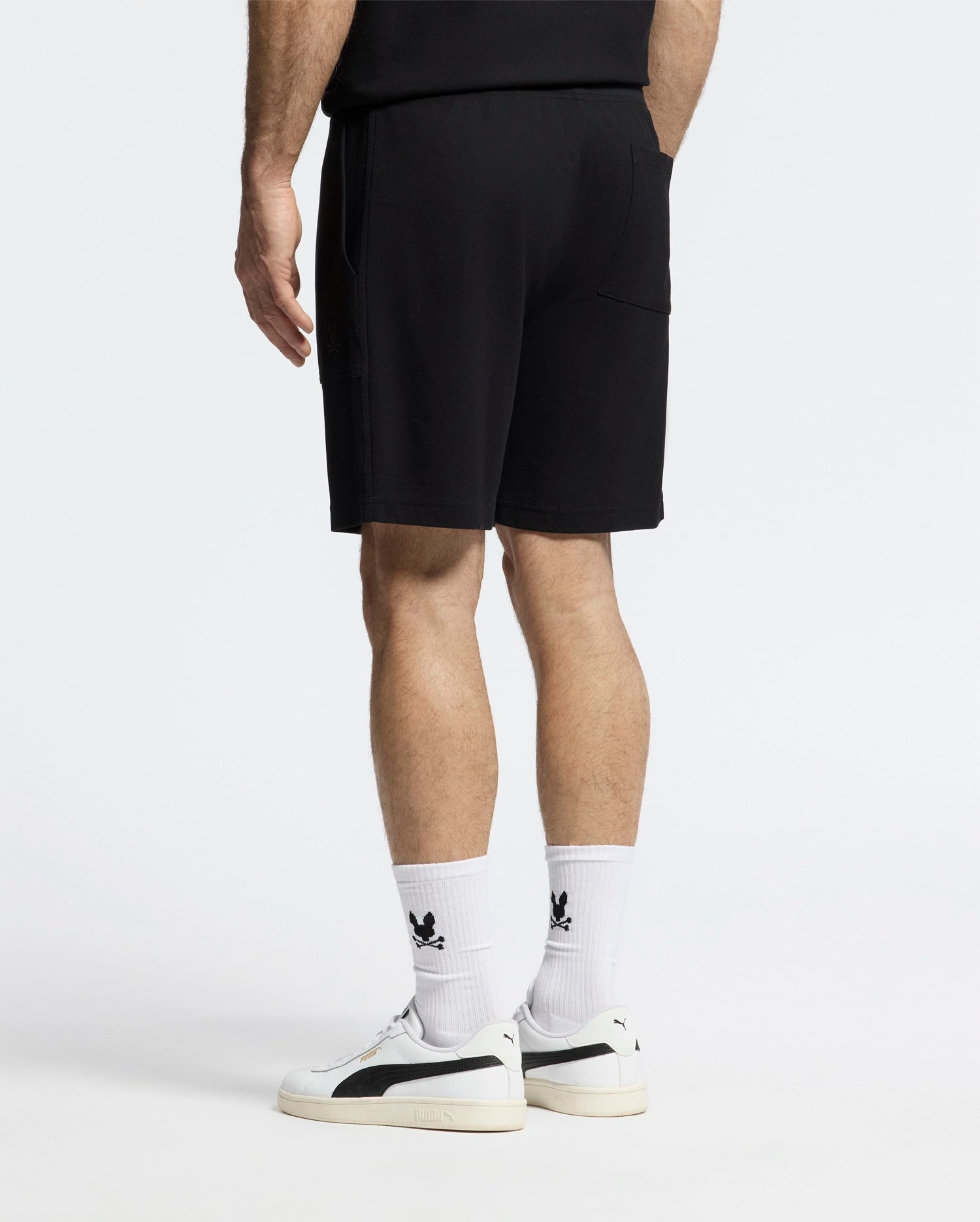 Dressed in a lightweight, black Pima cotton t-shirt and MENS TAYLOR LIGHT PIQUE SWEATSHORT by Psycho Bunny, they pair their outfit with white socks accented by a black logo and chic white sneakers featuring black details against a plain white backdrop, exuding effortless casual-chic style.
