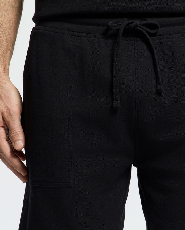 Close-up of someone in Psycho Bunny's black MENS TAYLOR LIGHT PIQUE SWEATSHORT (B6R344F200) and a black shirt, ideal for a casual-chic wardrobe. Made from lightweight Pima cotton, the sweatshorts feature an elastic waistband with drawstring. The left hand rests casually against a plain white backdrop.