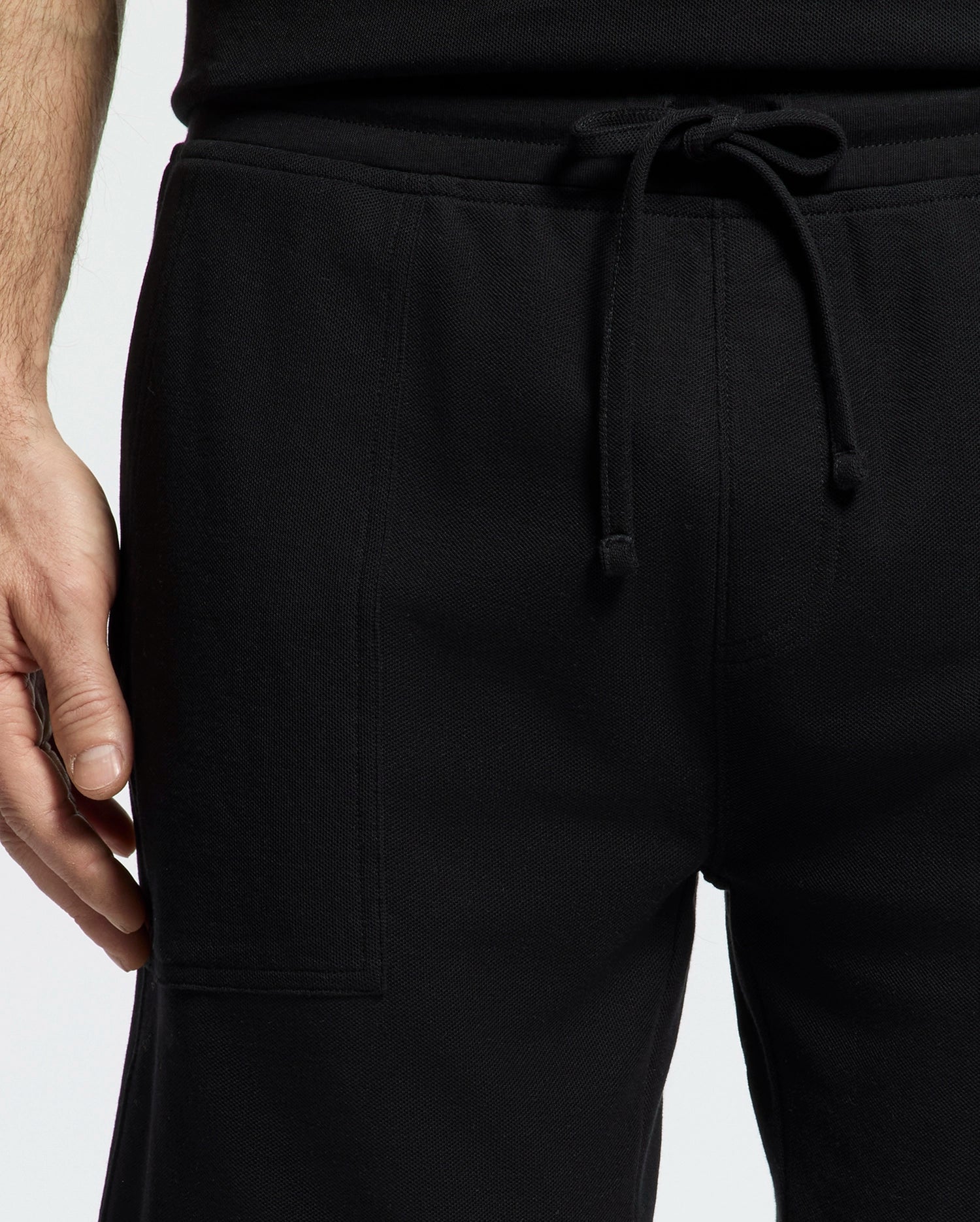 Close-up of someone in Psycho Bunny's black MENS TAYLOR LIGHT PIQUE SWEATSHORT (B6R344F200) and a black shirt, ideal for a casual-chic wardrobe. Made from lightweight Pima cotton, the sweatshorts feature an elastic waistband with drawstring. The left hand rests casually against a plain white backdrop.