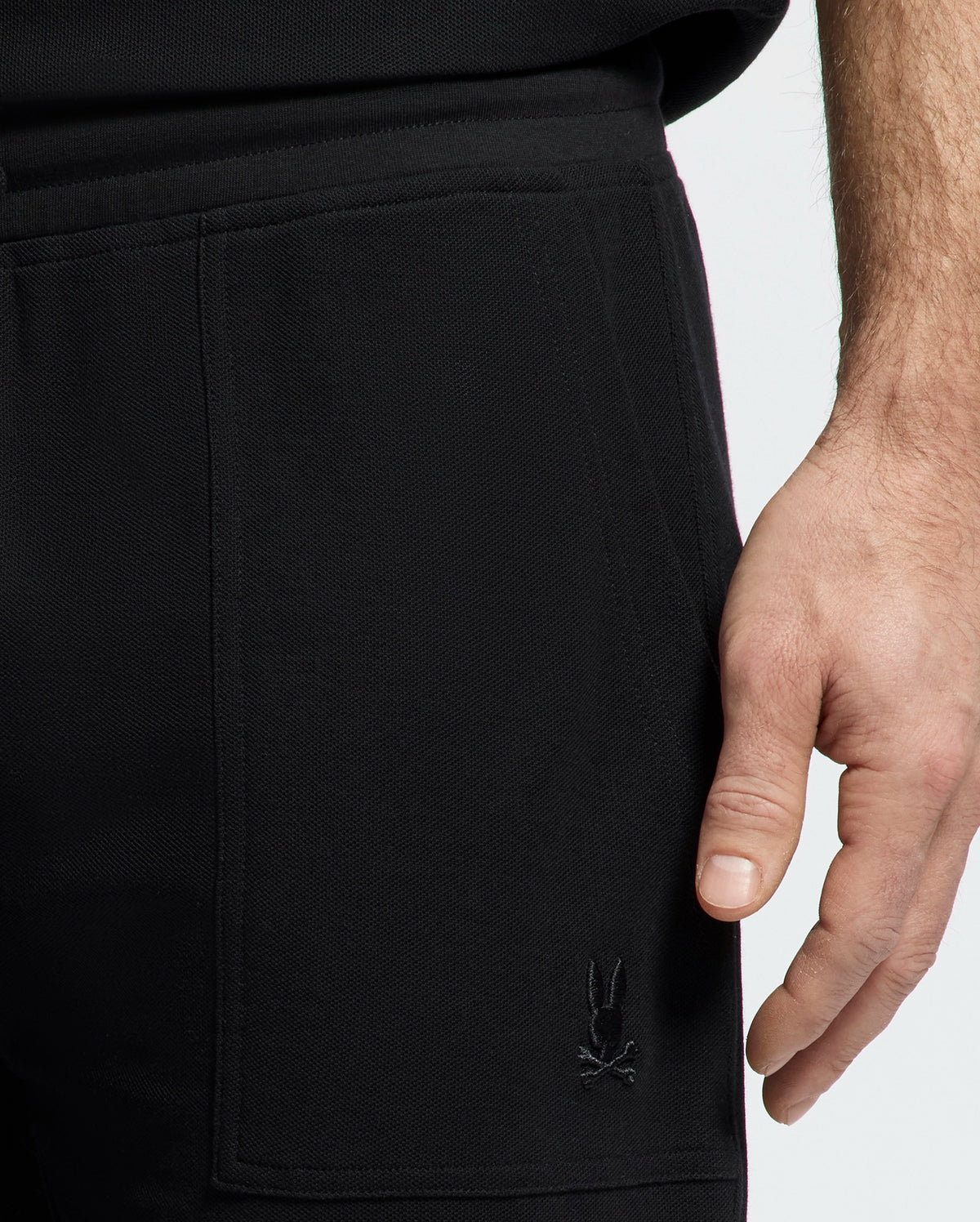 A person rests their hand on the side pocket of Psycho Bunny's MENS TAYLOR LIGHT PIQUE SWEATSHORT in black, crafted from lightweight Pima cotton with a small embroidered bunny skull logo near the pocket, against a plain background.