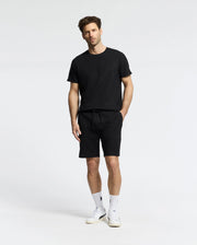 A man models in a Psycho Bunny black t-shirt and the MENS TAYLOR LIGHT PIQUE SWEATSHORT - B6R344F200 against a plain background, showcasing casual-chic style with lightweight fabrics. With short hair, beard, white socks, and sneakers, his look is complete.