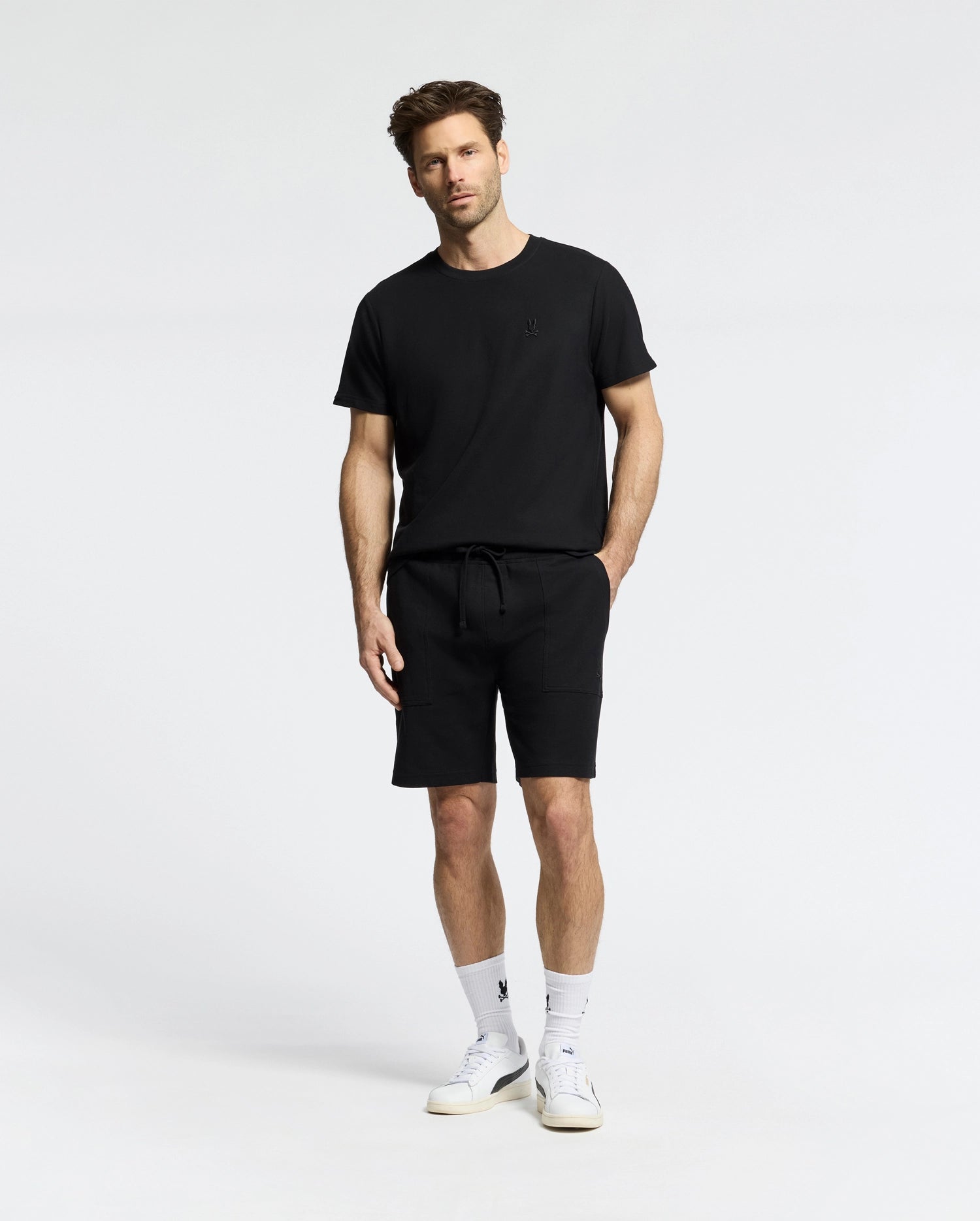 A man models in a Psycho Bunny black t-shirt and the MENS TAYLOR LIGHT PIQUE SWEATSHORT - B6R344F200 against a plain background, showcasing casual-chic style with lightweight fabrics. With short hair, beard, white socks, and sneakers, his look is complete.