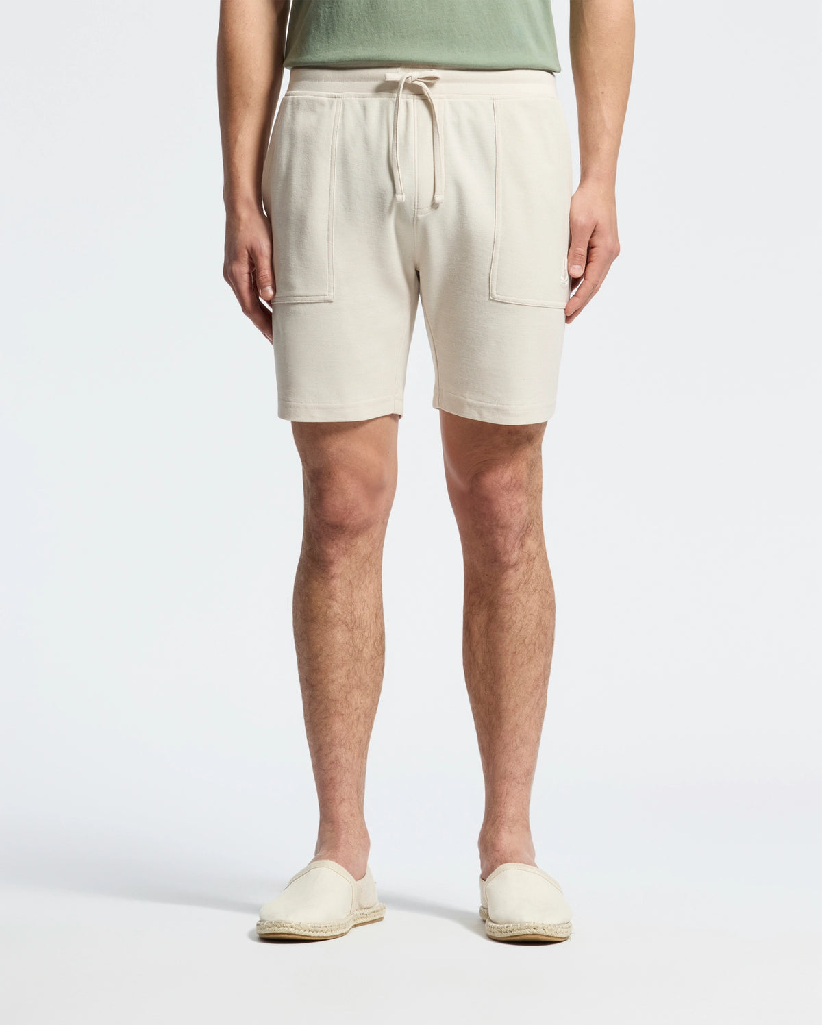 A person wearing Psycho Bunny's MENS TAYLOR LIGHT PIQUE SWEATSHORT in beige, featuring drawstrings and front pockets, paired with a green Pima cotton top and matched with beige slip-on shoes against a plain white background.