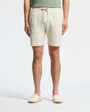 A person wearing Psycho Bunny's MENS TAYLOR LIGHT PIQUE SWEATSHORT in beige, featuring drawstrings and front pockets, paired with a green Pima cotton top and matched with beige slip-on shoes against a plain white background.