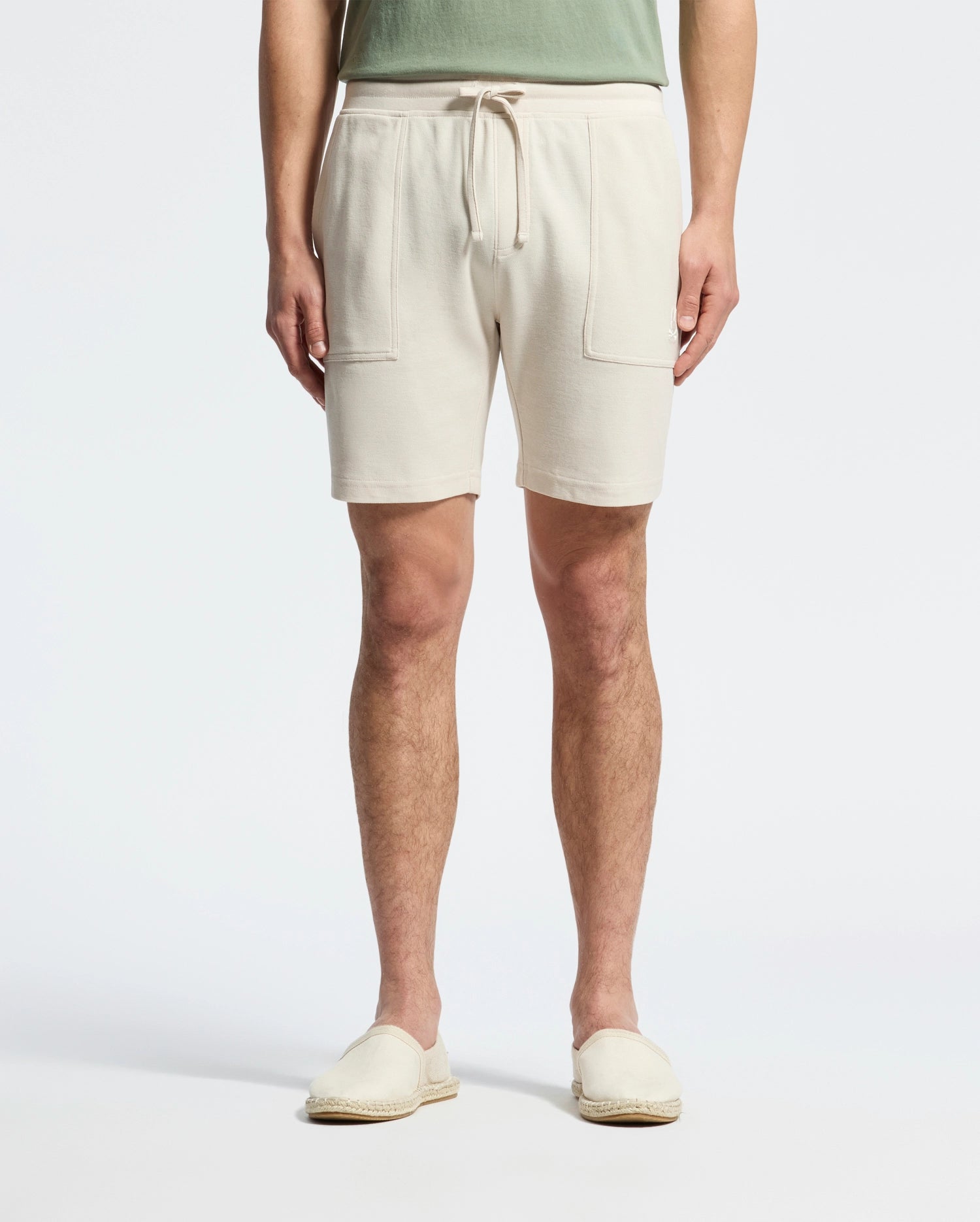 A person wearing Psycho Bunny's MENS TAYLOR LIGHT PIQUE SWEATSHORT in beige, featuring drawstrings and front pockets, paired with a green Pima cotton top and matched with beige slip-on shoes against a plain white background.