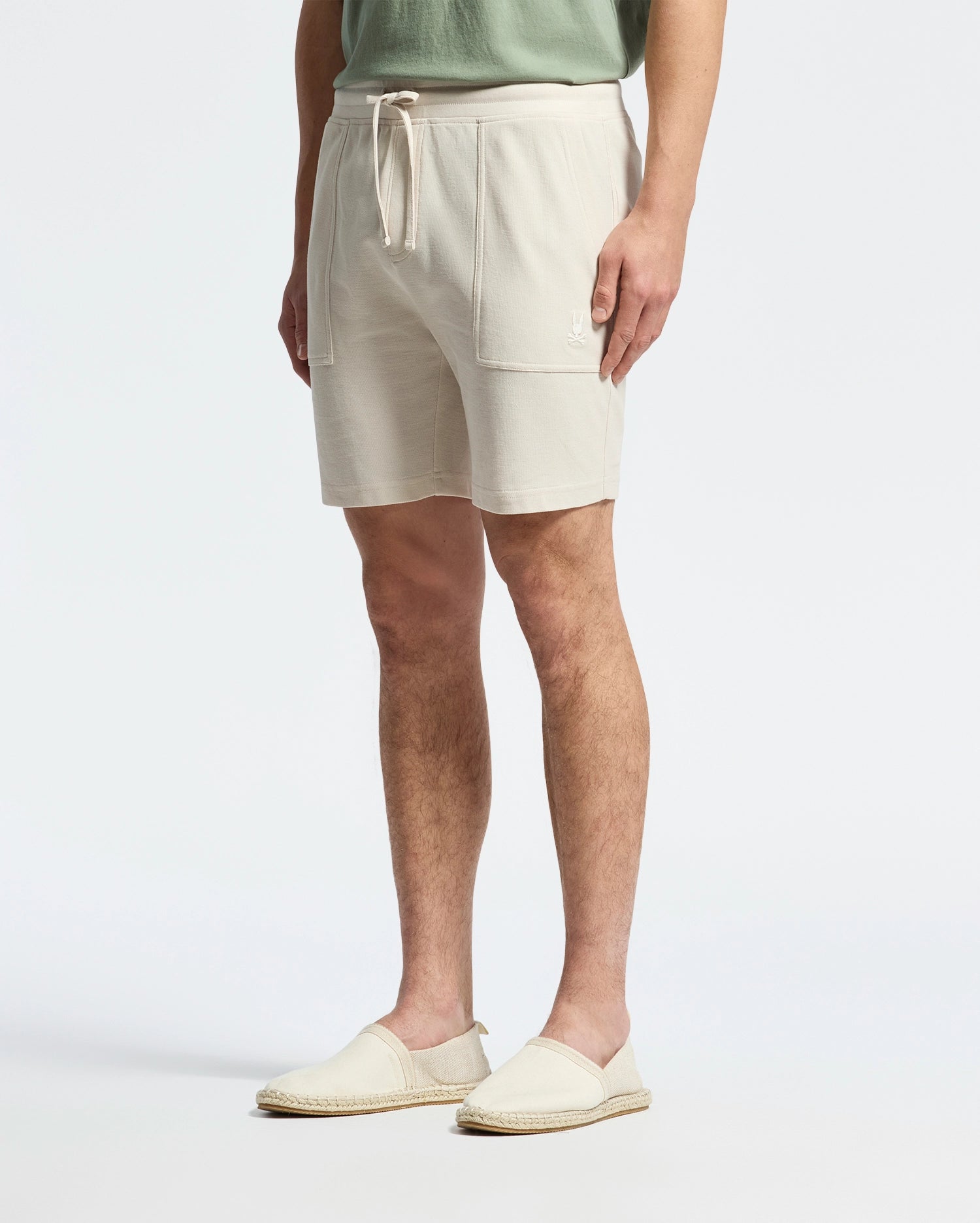 Sporting a casual-chic vibe in Psycho Bunny's MENS TAYLOR LIGHT PIQUE SWEATSHORT (B6R344F200), featuring pockets and a drawstring, paired with a lightweight light green shirt and beige espadrilles, they effortlessly stand against a plain white backdrop.