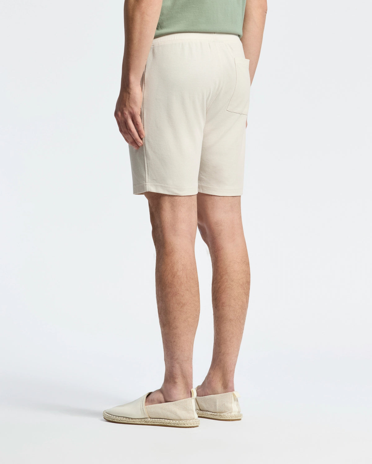 In front of a plain white background, a person turns away, sporting a casual-chic look with a lightweight light green Pima cotton shirt, Psycho Bunny's MENS TAYLOR LIGHT PIQUE SWEATSHORT in beige, and cream-colored slip-on shoes.