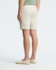 In front of a plain white background, a person turns away, sporting a casual-chic look with a lightweight light green Pima cotton shirt, Psycho Bunny's MENS TAYLOR LIGHT PIQUE SWEATSHORT in beige, and cream-colored slip-on shoes.