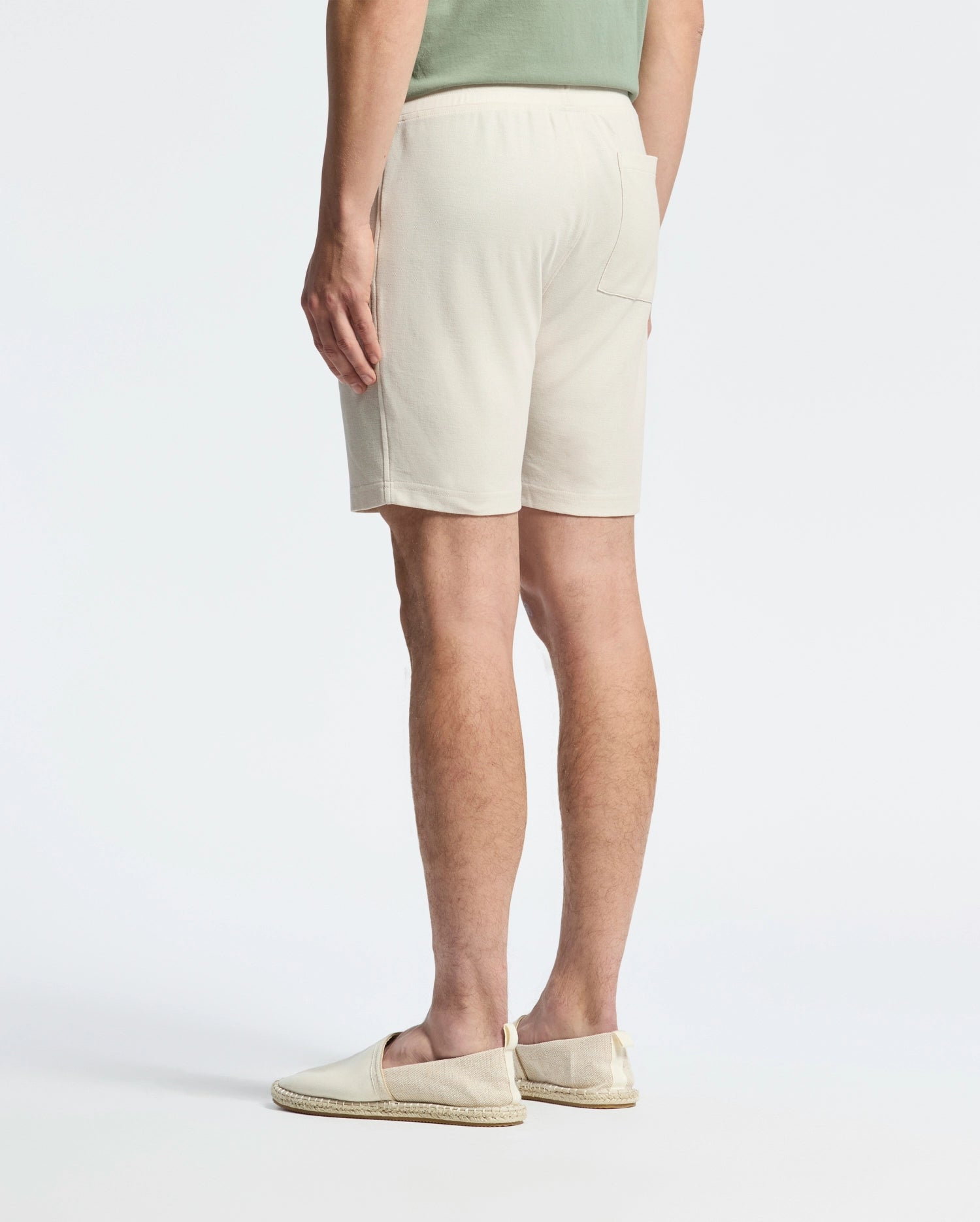 In front of a plain white background, a person turns away, sporting a casual-chic look with a lightweight light green Pima cotton shirt, Psycho Bunny's MENS TAYLOR LIGHT PIQUE SWEATSHORT in beige, and cream-colored slip-on shoes.