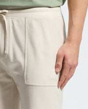 A person wears MENS TAYLOR LIGHT PIQUE SWEATSHORT - B6R344F200 by Psycho Bunny, lightweight beige with drawstrings and an embroidered rabbit's head logo on the pocket. They pair it casually chic with a green top, resting a hand near the pocket.