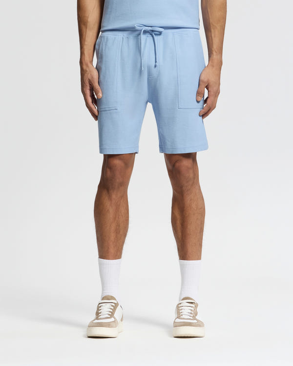 A person wearing Psycho Bunny's MENS TAYLOR LIGHT PIQUE SWEATSHORT in light blue, paired with white socks and white sneakers with beige accents, stands against a plain white background, epitomizing effortless casual-chic style.