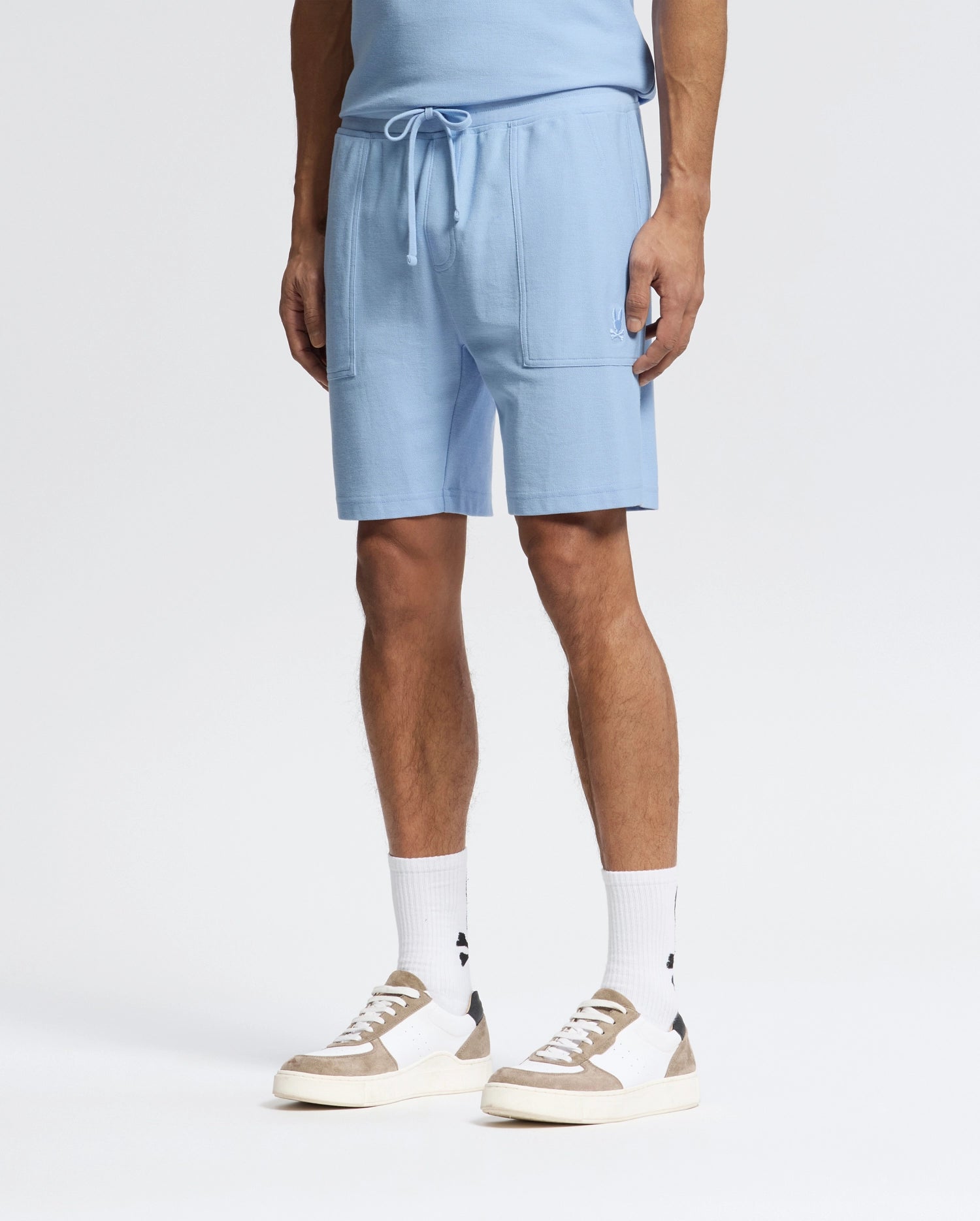 Wearing Psycho Bunny's MENS TAYLOR LIGHT PIQUE SWEATSHORT in Pima cotton, paired with a matching light blue shirt, white socks, and white sneakers with gray accents against a light gray backdrop creates a casual-chic look centered on the lower half.