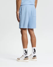 Wearing Psycho Bunny's Taylor Light Pique Sweatshorts in light blue, a coordinated shirt, white socks with black designs, and beige-white sneakers, the individual presents an effortlessly stylish look against a plain white background—ideal for a casual-chic wardrobe.