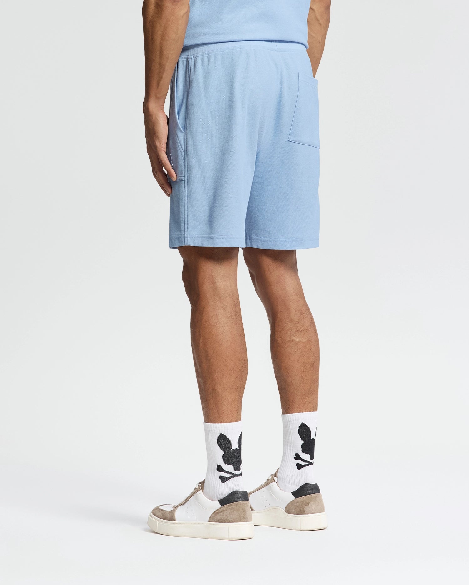 Wearing Psycho Bunny's Taylor Light Pique Sweatshorts in light blue, a coordinated shirt, white socks with black designs, and beige-white sneakers, the individual presents an effortlessly stylish look against a plain white background—ideal for a casual-chic wardrobe.