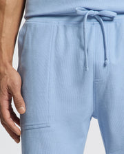 A person wearing Psycho Bunny's MENS TAYLOR LIGHT PIQUE SWEATSHORT in light blue Pima cotton with a drawstring waist and front pocket. Their left hand rests by their side against a plain white background, enhancing the casual-chic style.