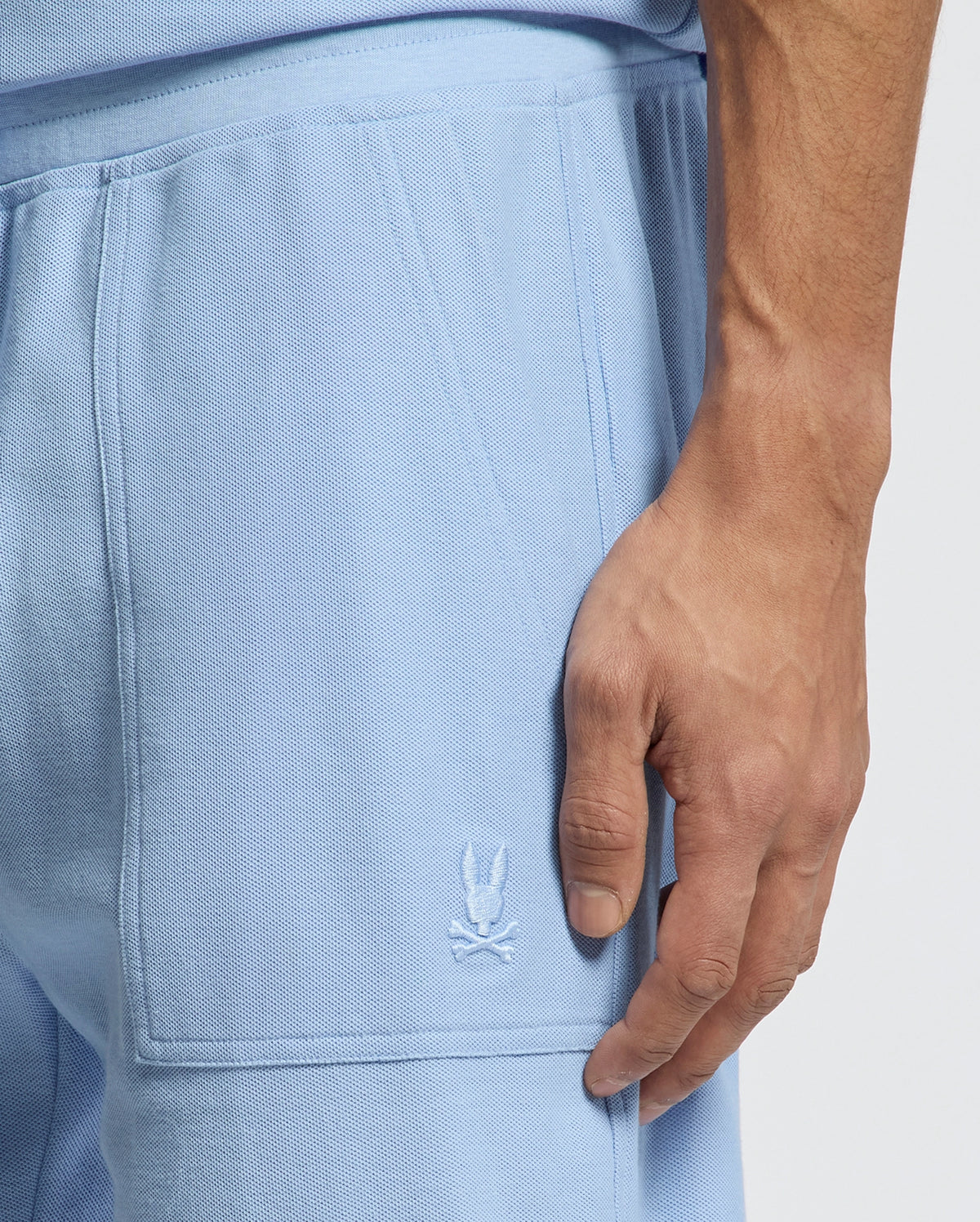 A close-up shows a person wearing Psycho Bunny's MENS TAYLOR LIGHT PIQUE SWEATSHORT in light blue, featuring a skull and crossed batons logo embroidered on the side. Made from Pima cotton, this textured fabric enhances a casual-chic vibe. The hand is near the pocket.