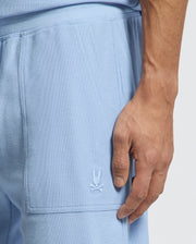A close-up shows a person wearing Psycho Bunny's MENS TAYLOR LIGHT PIQUE SWEATSHORT in light blue, featuring a skull and crossed batons logo embroidered on the side. Made from Pima cotton, this textured fabric enhances a casual-chic vibe. The hand is near the pocket.