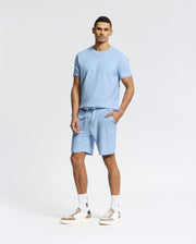 A man exudes casual-chic style against a plain backdrop, wearing a Psycho Bunny light blue Pima cotton tee with matching MENS TAYLOR LIGHT PIQUE SWEATSHORT - B6R344F200. He pairs this ensemble with white sneakers and socks, hands in his pockets, gazing directly at the camera.