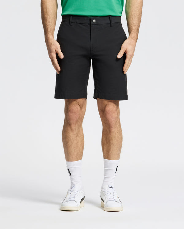 Against a plain backdrop, someone is wearing Psycho Bunny MENS FRANKLIN GOLF SHORT - B6R466F200 in black, a green shirt, white socks, and sneakers. The focus is on their lower half, highlighting the shorts' casual style.