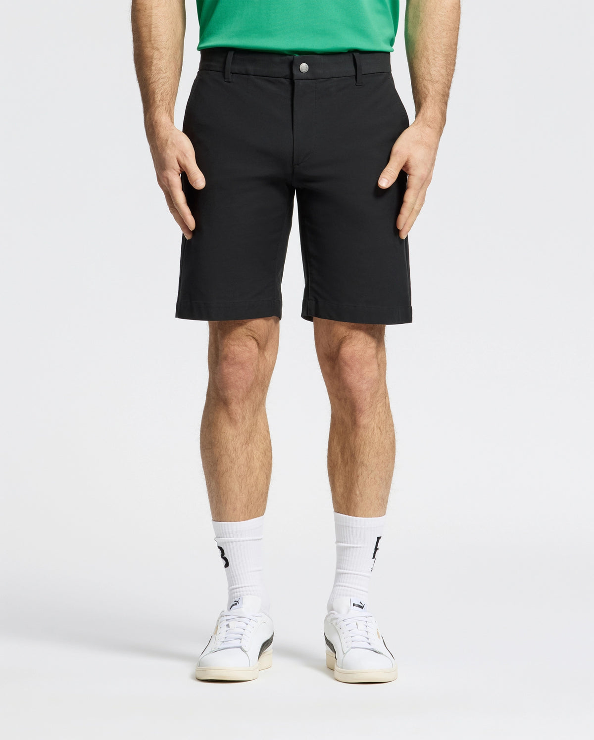Against a plain backdrop, someone is wearing Psycho Bunny MENS FRANKLIN GOLF SHORT - B6R466F200 in black, a green shirt, white socks, and sneakers. The focus is on their lower half, highlighting the shorts' casual style.