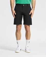 Against a plain backdrop, someone is wearing Psycho Bunny MENS FRANKLIN GOLF SHORT - B6R466F200 in black, a green shirt, white socks, and sneakers. The focus is on their lower half, highlighting the shorts' casual style.